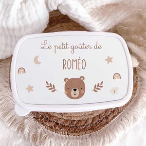 May include: A white rectangular lunch box featuring the text Le petit go&ucirc;ter de ROM&Eacute;O in brown lettering. The design includes a brown bear face, a crescent moon, stars, rainbows, leaves, and a butterfly in soft brown and beige tones.
