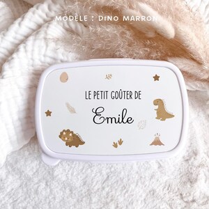 May include: A white rectangular lunch box featuring the text "LE PETIT GO&Ucirc;TER DE Emile" in black lettering. The lid is decorated with brown dinosaur illustrations, a volcano, stars, leaves, and footprints, with "MOD&Egrave;LE : DINO MARRON" printed above.