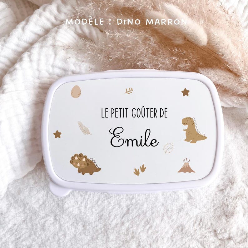 May include: A white rectangular lunch box featuring the text "LE PETIT GO&Ucirc;TER DE Emile" in black lettering. The lid is decorated with brown dinosaur illustrations, a volcano, stars, leaves, and footprints, with "MOD&Egrave;LE : DINO MARRON" printed above.