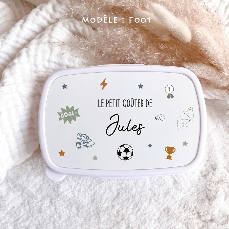 May include: A white rectangular lunch box with a lid featuring the text "LE PETIT GO&Ucirc;TER DE Jules" and various soccer-themed illustrations including a soccer ball, cleats, a trophy, a whistle, a goal sign, stars, and a lightning bolt.
