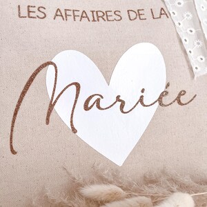 May include: A beige canvas tote bag with the text "LES AFFAIRES DE LA MARI&Eacute;E" in brown glitter lettering. A white heart is centered on the bag.