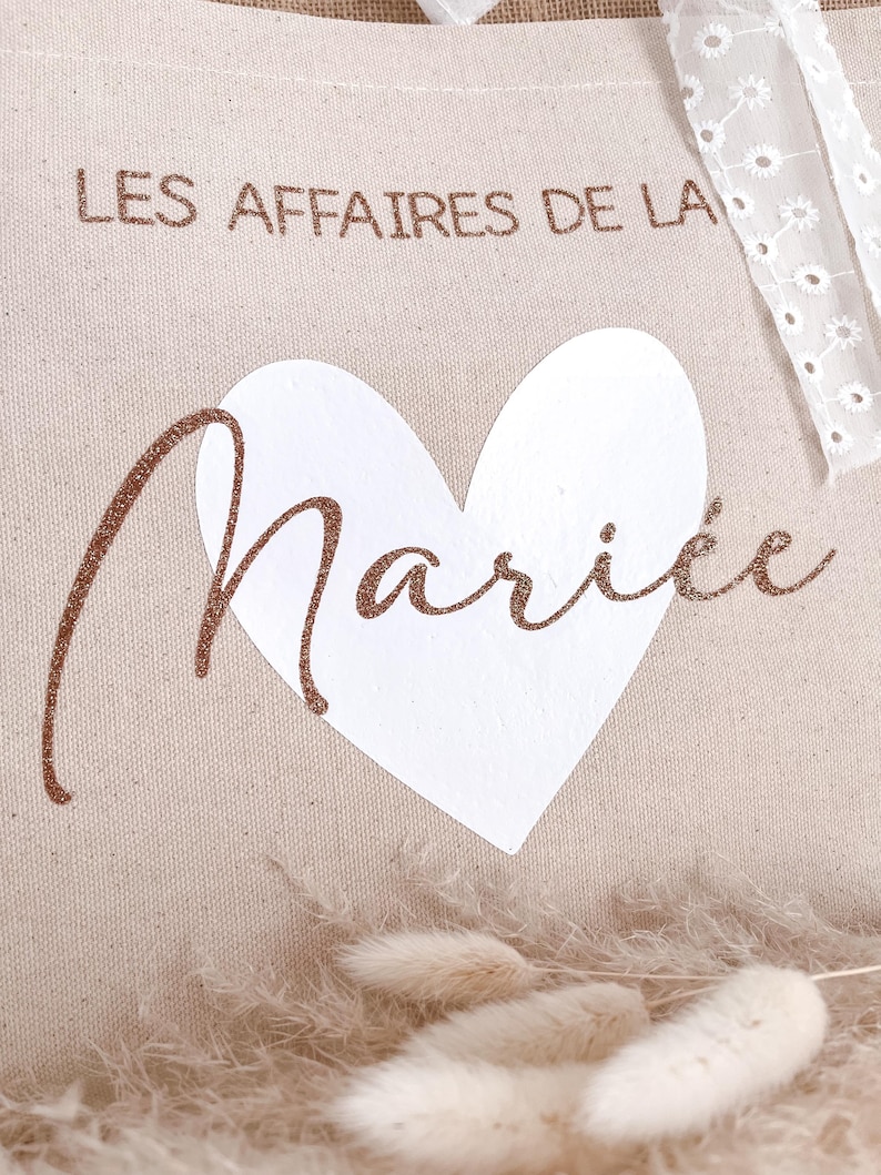 May include: A beige canvas tote bag with the text "LES AFFAIRES DE LA MARI&Eacute;E" in brown glitter lettering. A white heart is centered on the bag.