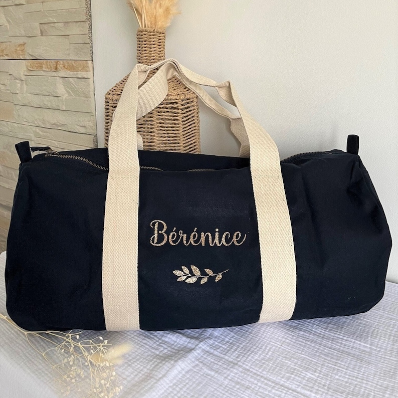 May include: A navy blue duffel bag with cream-colored handles and a zipper closure. The bag features the name "B&eacute;r&eacute;nice" embroidered in gold script, along with a gold leaf design. The bag is made of a sturdy fabric.