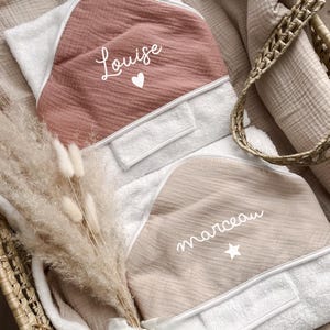 Op de afbeelding: Twee gepersonaliseerde babyhanddoeken met capuchon. Eén is stoffig roze met de naam "Louise" en een hart, de andere is beige met de naam "marceau" en een ster. Beide hebben witte randen. Op een neutrale achtergrond.