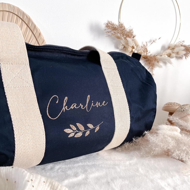 May include: Navy blue duffel bag with cream-colored straps. The bag features the name "Charline" in a cursive font, along with a gold glitter leaf design. The bag is made of a soft material.