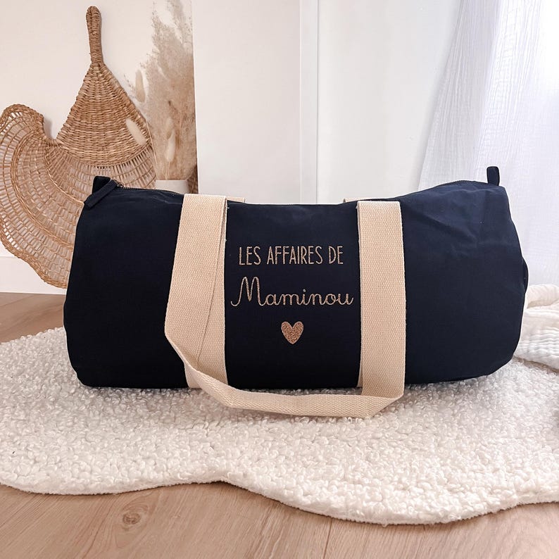 May include: A navy blue duffel bag with beige handles and the text "LES AFFAIRES DE Maminou" in gold script, along with a gold heart. The bag is cylindrical and sits on a white, fluffy surface.