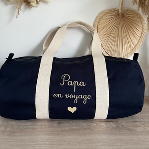 May include: Navy blue duffel bag with cream-colored handles and accents. The bag features the text "Papa en voyage" in gold script, along with a gold heart. The bag is designed for travel.