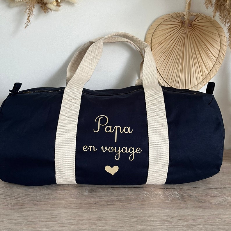 May include: Navy blue duffel bag with cream-colored handles and accents. The bag features the text "Papa en voyage" in gold script, along with a gold heart. The bag is designed for travel.
