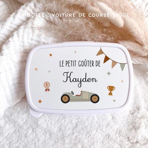 May include: A white rectangular lunch box featuring a sage green race car graphic, a gold trophy, a number one medal, and bunting. The text reads LE PETIT GO&Ucirc;TER DE Kayden. The design includes small star and plus sign accents in muted earth tones.