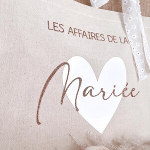 May include: A beige canvas tote bag with a white heart and the text "LES AFFAIRES DE LA MARI&Eacute;E" in rose gold glitter.
