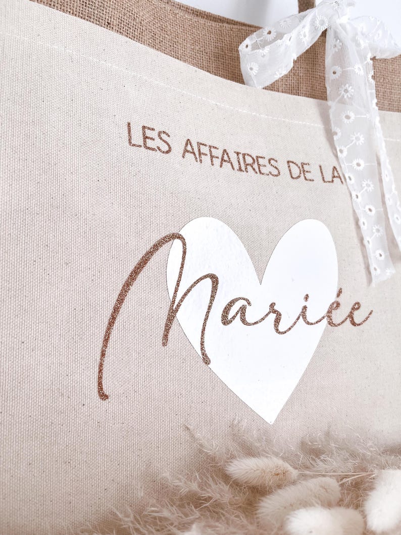 May include: A beige canvas tote bag with a white heart and the text "LES AFFAIRES DE LA MARI&Eacute;E" in rose gold glitter.