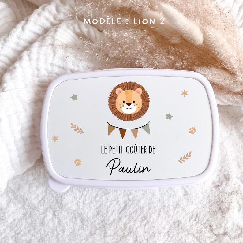 May include: A white rectangular lunch box featuring a cartoon lion illustration, a bunting banner, and the text "MOD&Egrave;LE : LION 2", "LE PETIT GO&Ucirc;TER DE", and "Paulin". The design includes small stars, paw prints, and leaf motifs in muted earth tones.