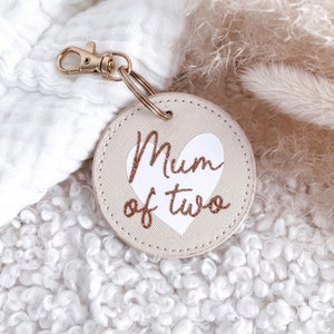 Peut inclure: Un porte-clés rond beige avec un cœur blanc et le texte "Mum of two" en lettres pailletées dorées.