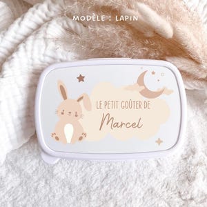 May include: A white rectangular lunch box featuring a beige cartoon rabbit, a crescent moon, stars, and clouds. The text reads "MOD&Egrave;LE : LAPIN" at the top and "LE PETIT GO&Ucirc;TER DE Marcel" in the center, printed in a brown script and sans-serif font.