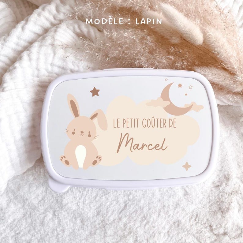 May include: A white rectangular lunch box featuring a beige cartoon rabbit, a crescent moon, stars, and clouds. The text reads "MOD&Egrave;LE : LAPIN" at the top and "LE PETIT GO&Ucirc;TER DE Marcel" in the center, printed in a brown script and sans-serif font.