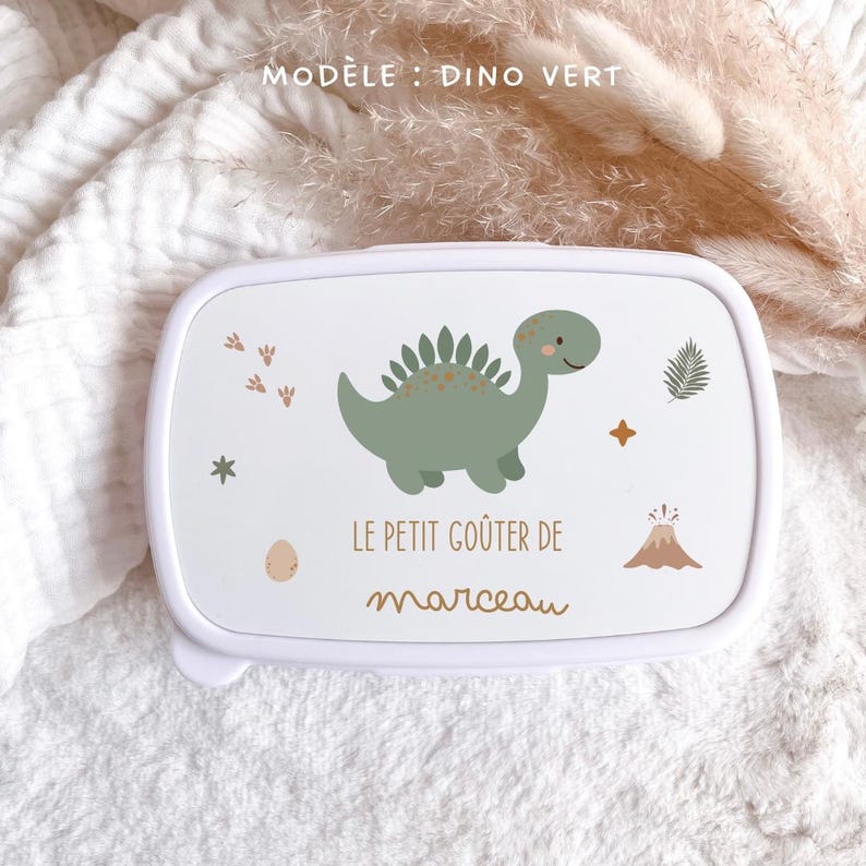 May include: A white rectangular snack box featuring a green dinosaur illustration, a small volcano, a fern leaf, and footprints. The text reads "LE PETIT GO&Ucirc;TER DE marceau" in brown lettering. The lid is white with rounded corners and a smooth finish.