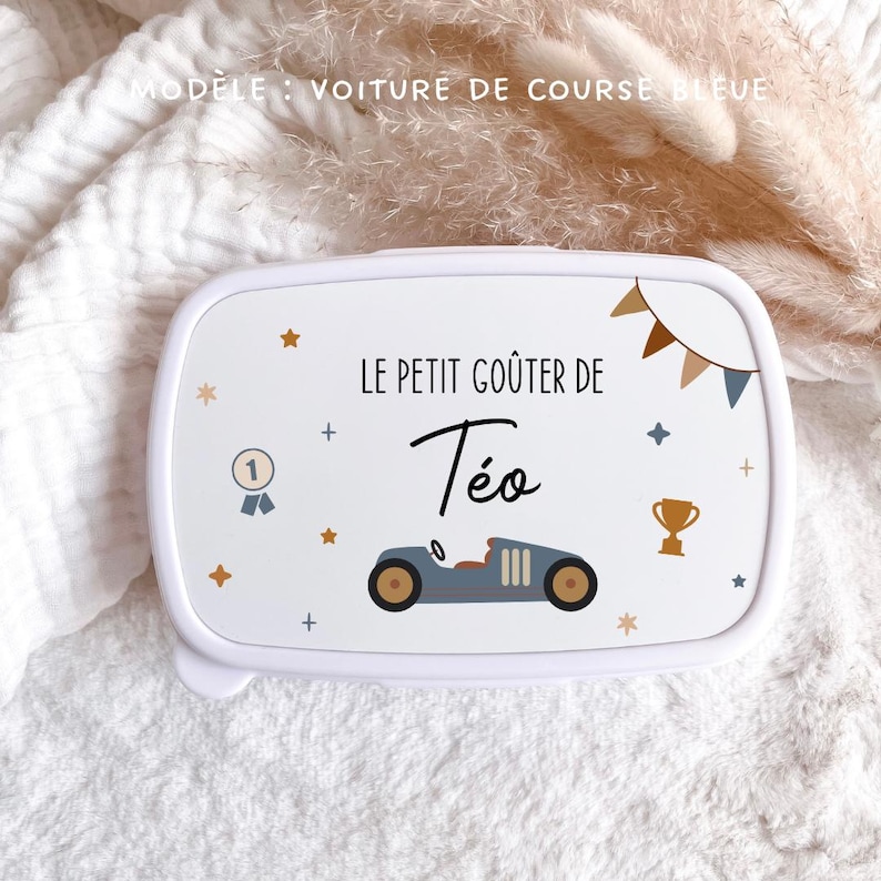 May include: A white rectangular lunch box featuring the text LE PETIT GO&Ucirc;TER DE T&eacute;o, a blue vintage race car illustration, a gold trophy, a number one medal, and small star icons. The lid also displays a decorative bunting banner design.
