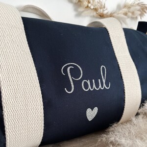 May include: Navy blue duffel bag with cream-colored straps. The name "Paul" is printed in silver glitter script, with a silver glitter heart below. The bag is made of a sturdy fabric and is designed for travel or gym use.