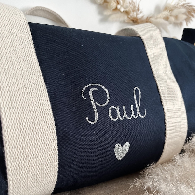 May include: Navy blue duffel bag with cream-colored straps. The name "Paul" is printed in silver glitter script, with a silver glitter heart below. The bag is made of a sturdy fabric and is designed for travel or gym use.