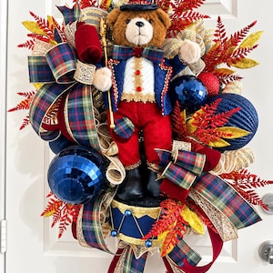 May include: A decorative wreath featuring a plush brown bear dressed as a ringmaster in a blue jacket, red pants, and top hat. The wreath is adorned with red and gold fern sprigs, plaid ribbons, and large blue and red spherical ornaments.