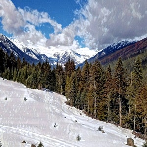 Può includere: Un paesaggio montano innevato con una foresta di alberi sempreverdi in primo piano. Il cielo è blu con nuvole bianche.
