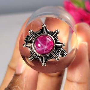 May include: A silver-coloured ring featuring a starburst design and a large, round, pink gemstone. The ring is set on a clear, cylindrical object. The gemstone has a star-like pattern within it.