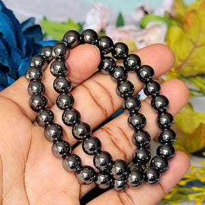 Peut inclure: Deux bracelets de perles d'hématite. Chaque bracelet est composé de perles sphériques polies, gris foncé. Les bracelets sont tenus dans la main d'une personne, avec un arrière-plan flou de fleurs.