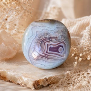 Puede incluir: Una piedra de &aacute;gata pulida y redondeada con intrincadas bandas conc&eacute;ntricas en tonos azul p&aacute;lido, blanco y lavanda suave. La superficie es lisa y reflectante, mostrando un patr&oacute;n natural en capas que irradia desde un punto central.