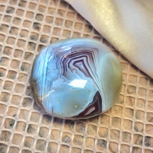 Puede incluir: Una piedra de &aacute;gata pulida y redondeada que presenta intrincadas bandas conc&eacute;ntricas en tonos azul claro, blanco y marr&oacute;n rojizo intenso. La superficie es lisa y reflectante, mostrando capas minerales naturales y un aspecto v&iacute;treo.