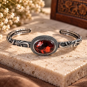 May include: An open cuff bracelet made of oxidized silver featuring an oval centerpiece with a deep red resin inlay. The resin contains a visible embedded red flower design. The silver band is decorated with intricate scrollwork and rope detailing.