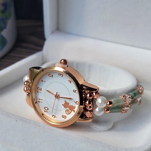 Op de afbeelding: Roségouden katvormig horloge met een witte wijzerplaat, met een katten silhouet. De band is versierd met groene kralen, parels en roségouden accenten. Het horloge wordt gepresenteerd in een witte doos.