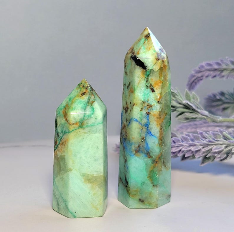 May include: Two polished crystal towers with a hexagonal base and pointed top. The stones feature a light green base color with prominent veins of brown, tan, and bright blue mineral inclusions throughout the smooth, semi-translucent surfaces.