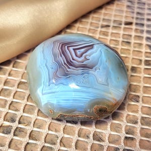 Puede incluir: Una piedra de &aacute;gata pulida y redondeada que presenta bandas conc&eacute;ntricas intrincadas en tonos azul claro, blanco y violeta intenso. La superficie es lisa y brillante, mostrando capas geol&oacute;gicas naturales y una peque&ntilde;a inclusi&oacute;n marr&oacute;n texturizada.