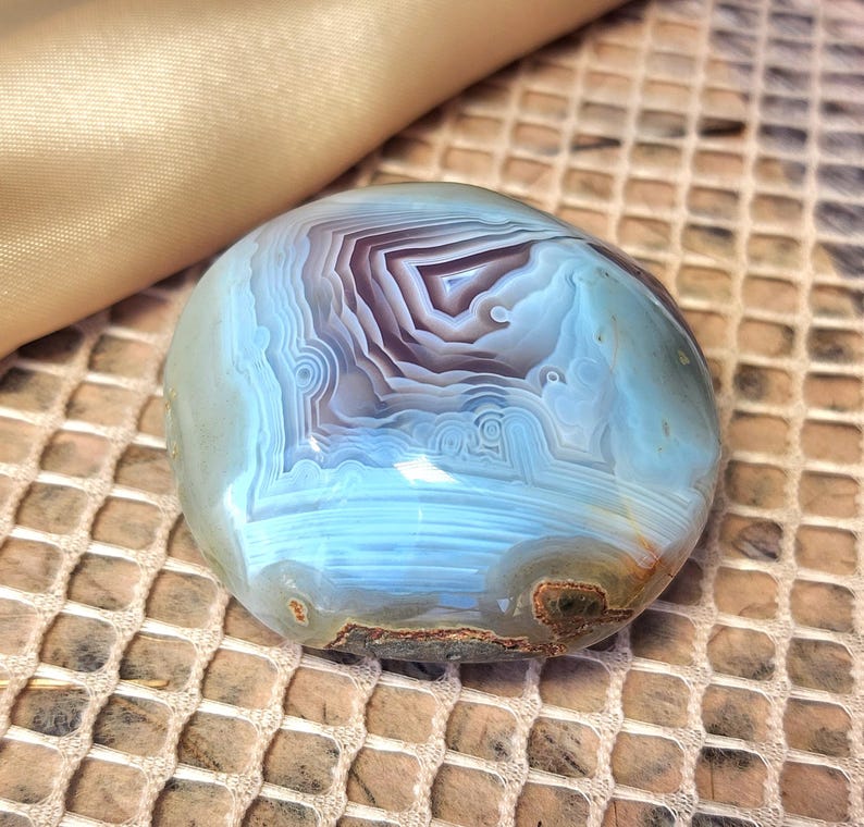 Puede incluir: Una piedra de &aacute;gata pulida y redondeada que presenta bandas conc&eacute;ntricas intrincadas en tonos azul claro, blanco y violeta intenso. La superficie es lisa y brillante, mostrando capas geol&oacute;gicas naturales y una peque&ntilde;a inclusi&oacute;n marr&oacute;n texturizada.