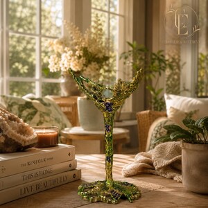 May include: A decorative green metal goblet featuring a textured vine and leaf design with small purple accents along the stem and base. The object stands upright on a flat surface, showcasing an intricate, organic structure with a wide, open bowl.