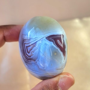 Puede incluir: Una piedra preciosa de &aacute;gata pulida de forma ovalada con intrincadas bandas conc&eacute;ntricas en tonos azul claro transl&uacute;cido, morado intenso y blanco. La piedra tiene una superficie lisa y brillante que refleja la luz, resaltando las capas minerales.