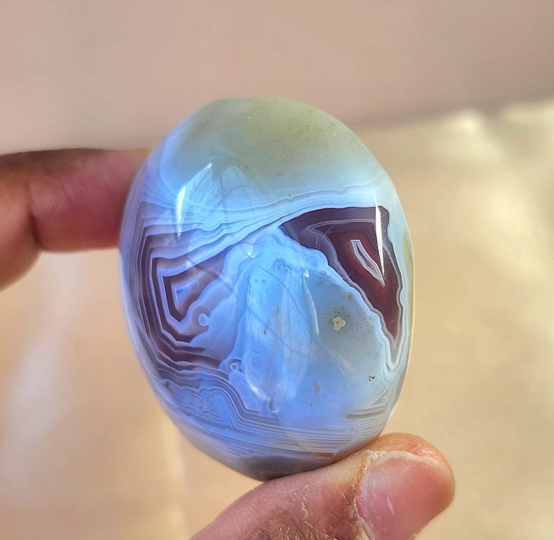 Puede incluir: Una piedra preciosa de &aacute;gata pulida de forma ovalada con intrincadas bandas conc&eacute;ntricas en tonos azul claro transl&uacute;cido, morado intenso y blanco. La piedra tiene una superficie lisa y brillante que refleja la luz, resaltando las capas minerales.