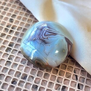 Puede incluir: Una piedra de &aacute;gata pulida y redondeada que presenta intrincadas bandas conc&eacute;ntricas en tonos azul p&aacute;lido, gris suave y marr&oacute;n tierra. La superficie es lisa y reflectante, destacando los patrones minerales naturales en capas.