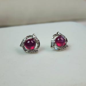K&ouml;nnte beinhalten: Ein Paar Ohrstecker mit einem zentralen, rund geschliffenen, tiefrosa Cabochon-Edelstein in einer silberfarbenen Metallfassung. Die Fassung ist mit kleinen, klaren, funkelnden Steinen in einem geschwungenen, offenen Design verziert.
