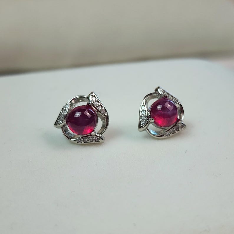 K&ouml;nnte beinhalten: Ein Paar Ohrstecker mit einem zentralen, rund geschliffenen, tiefrosa Cabochon-Edelstein in einer silberfarbenen Metallfassung. Die Fassung ist mit kleinen, klaren, funkelnden Steinen in einem geschwungenen, offenen Design verziert.
