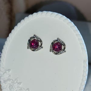 K&ouml;nnte beinhalten: Ein Paar Ohrstecker mit einem runden, tiefrosa Cabochon-Stein, der einen sechsstrahligen Sterneffekt aufweist. Die Steine sind in eine silberfarbene Metallfassung eingefasst, die mit kleinen, klaren, funkelnden Steinen in geschwungener Form verziert ist.