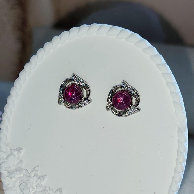 K&ouml;nnte beinhalten: Ein Paar Ohrstecker mit einem runden, tiefrosa Cabochon-Stein, der einen sechsstrahligen Sterneffekt aufweist. Die Steine sind in eine silberfarbene Metallfassung eingefasst, die mit kleinen, klaren, funkelnden Steinen in geschwungener Form verziert ist.