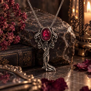 Op de afbeelding: Een zilverkleurige hanger aan een ketting met een centrale, ovaalvormige, dieprode edelsteen. De metalen zetting is vormgegeven als een staande figuur met vloeiende gewaden, hangend aan een fijne zilverkleurige ketting met vierkante schakels.