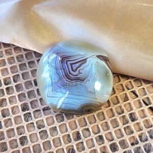 Puede incluir: Una piedra de &aacute;gata pulida y redondeada que presenta bandas conc&eacute;ntricas intrincadas en tonos azul claro, blanco y marr&oacute;n oscuro. La superficie es lisa y reflectante, mostrando capas geol&oacute;gicas naturales con un patr&oacute;n definido.