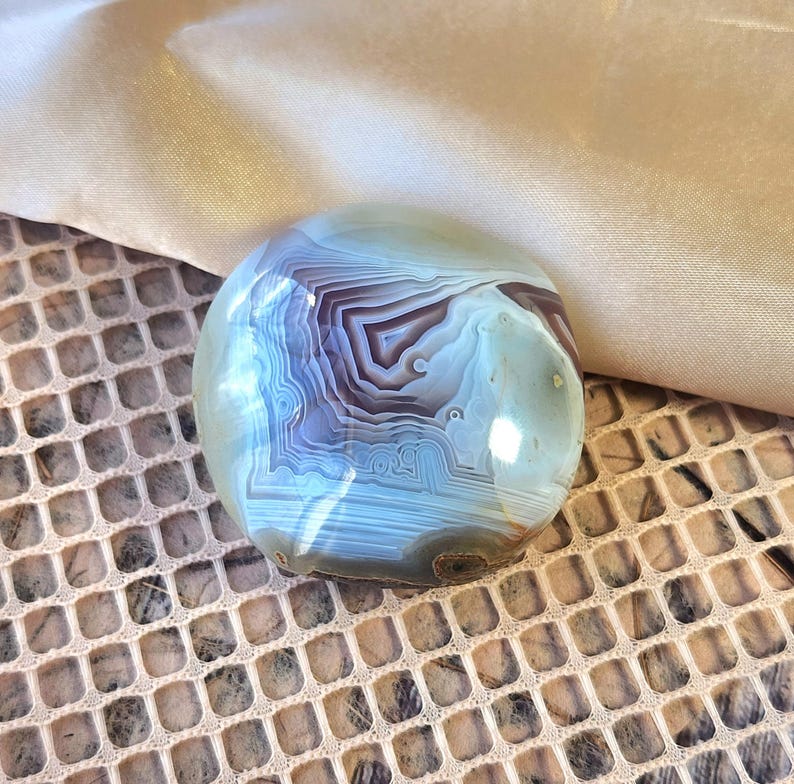 Puede incluir: Una piedra de &aacute;gata pulida y redondeada que presenta bandas conc&eacute;ntricas intrincadas en tonos azul claro, blanco y marr&oacute;n oscuro. La superficie es lisa y reflectante, mostrando capas geol&oacute;gicas naturales con un patr&oacute;n definido.