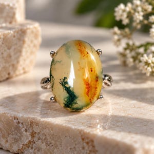 May include: A silver ring featuring a large, oval-shaped cabochon stone. The stone is translucent with a creamy off-white base, displaying distinct moss-like green inclusions and vibrant orange-red mineral patterns throughout the surface.