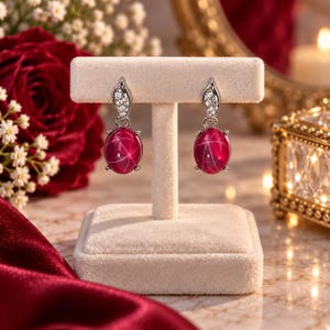 May include: A pair of drop earrings featuring oval-shaped, deep pink star gemstones suspended from silver-toned settings. Each earring includes a small, teardrop-shaped accent piece encrusted with clear, sparkling stones above the main gem.