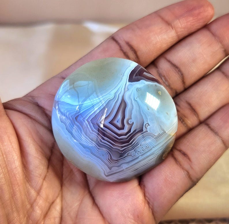 Puede incluir: Una piedra de &aacute;gata pulida y redondeada con intrincadas bandas conc&eacute;ntricas en tonos azul claro, blanco y morado intenso. La superficie es lisa y reflectante, mostrando un patr&oacute;n natural en capas que irradia desde el centro hacia afuera.