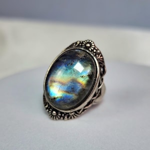 May include: An oval-shaped labradorite gemstone ring set in a decorative silver band. The stone displays iridescent flashes of blue, yellow, and green light across its polished surface. The metal setting features intricate floral-inspired detailing.