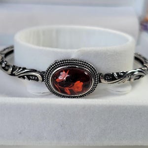 May include: Silver-toned bangle bracelet with an oval, dark red centerpiece featuring a floral design in shades of red and white. The bracelet has an ornate, scrolling design on the band and around the centerpiece.